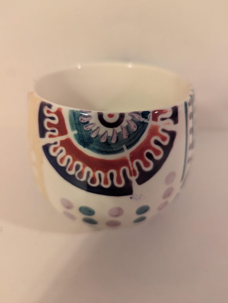 May include: A white ceramic cup with a colorful geometric design. The design features a large circle with a smaller circle inside, surrounded by a wavy line. The cup is decorated with dots and swirls in shades of blue, green, purple, and red.