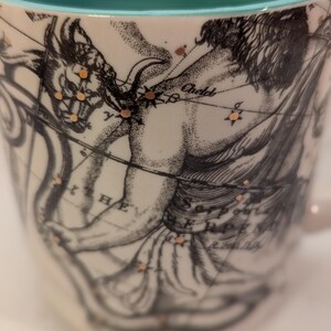 May include: A white ceramic mug with a turquoise rim. The mug features a black and white illustration of a constellation with gold accents. The constellation is labeled "Andromeda".