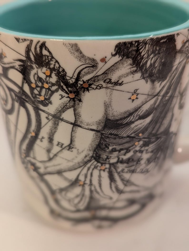 May include: A white ceramic mug with a turquoise rim. The mug features a black and white illustration of a constellation with gold accents. The constellation is labeled "Andromeda".