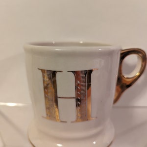 Pu&ograve; includere: Tazza in ceramica bianca con una lettera H dorata sul davanti. La tazza ha un bordo e un manico dorati.