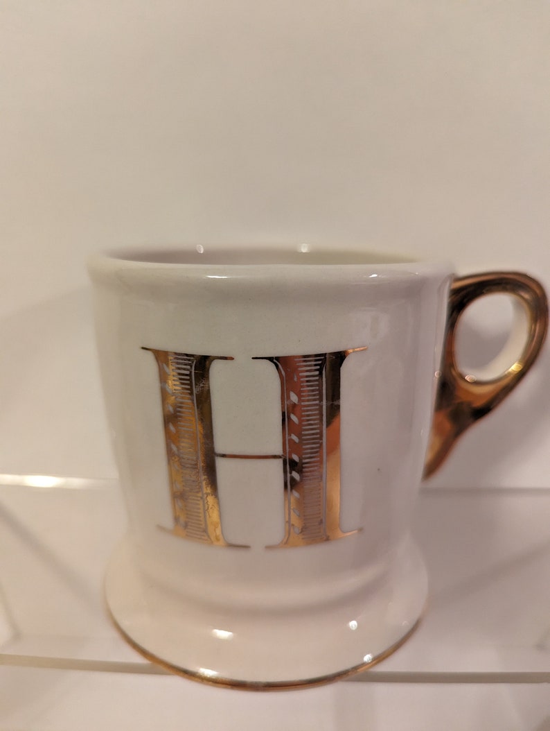 Pu&ograve; includere: Tazza in ceramica bianca con una lettera H dorata sul davanti. La tazza ha un bordo e un manico dorati.