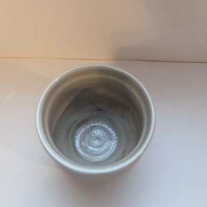 May include: A white ceramic cup with a slightly textured surface. The cup is empty and has a circular shape.