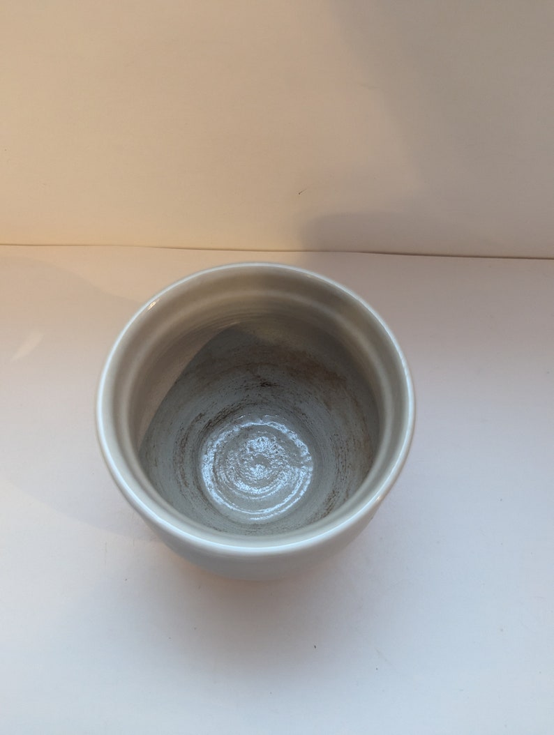 May include: A white ceramic cup with a slightly textured surface. The cup is empty and has a circular shape.