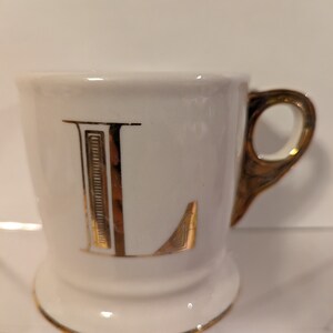 Pu&ograve; includere: Tazza in ceramica bianca con una lettera L dorata sul davanti. La tazza ha un bordo e un manico dorati.