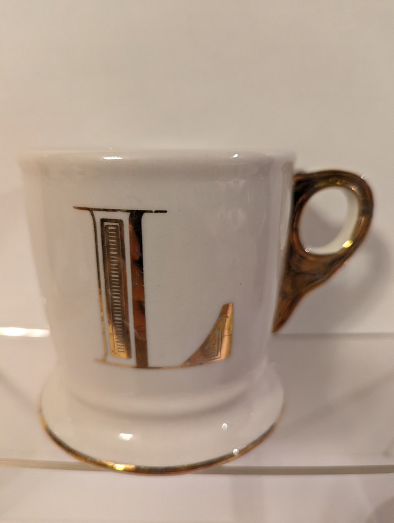 Pu&ograve; includere: Tazza in ceramica bianca con una lettera L dorata sul davanti. La tazza ha un bordo e un manico dorati.