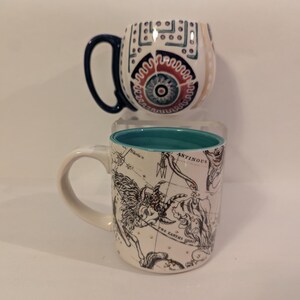 May include: Two ceramic mugs. The first mug has a white background with a blue, red, and white geometric design. The second mug has a white background with a black and white constellation design and a turquoise rim.