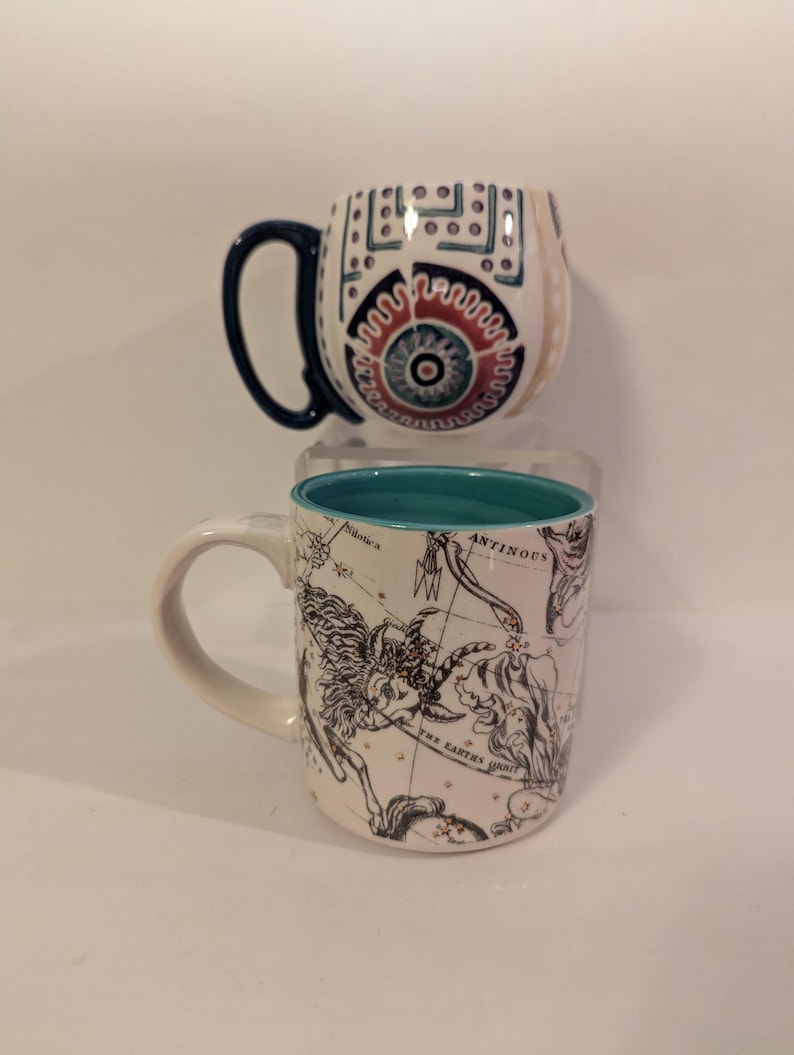 May include: Two ceramic mugs. The first mug has a white background with a blue, red, and white geometric design. The second mug has a white background with a black and white constellation design and a turquoise rim.