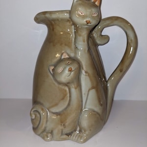 May include: A ceramic vase shaped like two cats, one sitting on top of the other. The vase is a light brown color with a glaze finish. The cats have brown eyes and a light brown nose.