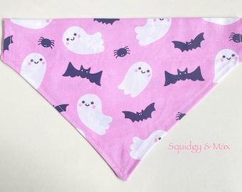 May include: Pink Halloween-themed pet bandana featuring a repeating pattern of white ghosts, dark purple bats, and spiders. The bandana is triangular in shape and made of fabric.