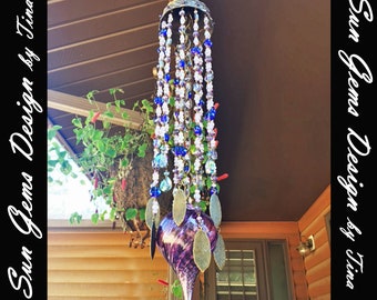 May include: A handcrafted wind chime with a decorative metal top and strands of colorful beads. The wind chime features a purple glass teardrop-shaped base with gold-colored leaf accents. The text "Sun Gems Design by Tina" is visible.