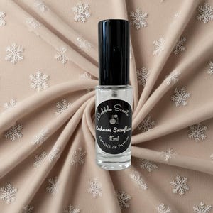 May include: A clear glass bottle of perfume with a black cap and label. The label reads "Dabble Scents Cashmere Snowflake 15ml Extrait de Parfum".