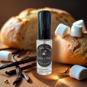 May include: A clear perfume bottle with a black cap labeled "Dabble Scents Mallo x Toast Extrait de Parfum" sits in front of a slice of bread, marshmallows, and vanilla beans. The bottle is the focal point, with the other items providing a warm, inviting backdrop.