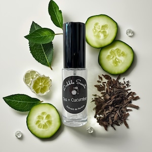 May include: A clear glass perfume bottle with a black cap, labeled "Tea + Cucumber" with "Extrait de Parfum." The bottle is surrounded by cucumber slices, green leaves, wood chips, and decorative crystals, suggesting a fresh, natural fragrance.