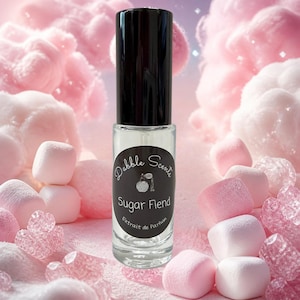 May include: A clear glass perfume roller bottle with a black cap and a black oval label. The label features the text Dabble Scents, an illustration of a perfume bottle, the words Sugar Fiend, and the phrase Extrait de Parfum in white text.