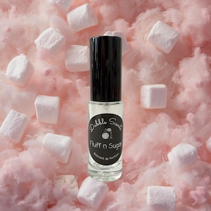 May include: A clear glass perfume roller bottle with a black cap and a black circular label. The label features the text Dabble Scents, an illustration of a perfume bottle, and the text Fluff n Sugar Extrait de Parfum in white lettering.