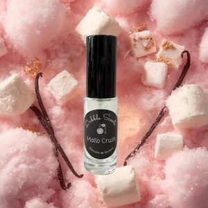 May include: A clear glass perfume roller bottle with a black cap and a black circular label featuring the text Dabble Scents Mallo Crush Extrait de Parfum. The bottle is surrounded by white marshmallows and dark brown vanilla bean pods.
