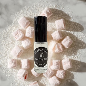 May include: A clear glass perfume roller bottle with a black cap and a black circular label. The label features the text Dabble Scents and Just The Fluff Extrait de Parfum with a small graphic of a perfume bottle. The bottle is surrounded by pink marshmallows.