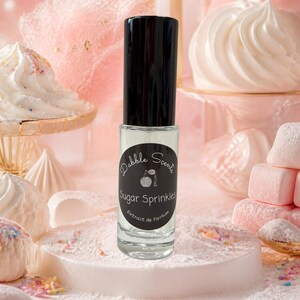 May include: A clear glass perfume bottle with a black cylindrical cap and a black circular label. The label features the white text Dabble Scents, Sugar Sprinkles, and Extrait de Parfum, along with a small graphic of a vintage perfume atomizer.