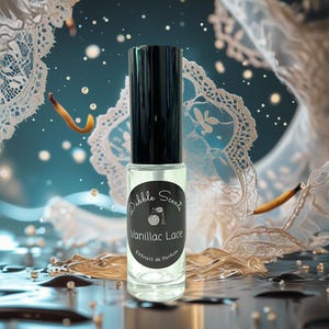 May include: A clear glass perfume bottle with a black cap. The bottle has a black label with the words "Dabble Scents" and "Vanillac Lace" printed on it. The background features lace and water droplets.