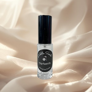 May include: A clear glass perfume bottle with a black cap. The bottle has a label with the words "Dabble Scents" and "Thirteenth" printed on it. The words "Extrait de Parfum" are also printed on the label. The background is a light beige fabric.
