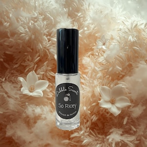 May include: A clear glass perfume bottle with a black cap, labeled "Dabble Scents So Ricey Extrait de Parfum." The bottle is set against a backdrop of white flowers and a textured, beige background.