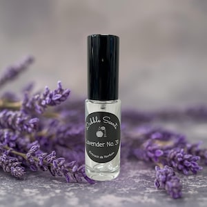 May include: A clear glass perfume bottle with a black cap, labeled "Dabble Scents Lavender No. 31 Extrait de Parfum." The bottle is surrounded by sprigs of purple lavender, set against a gray background.