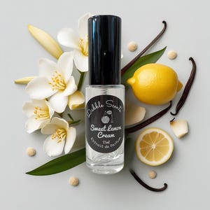 May include: A clear perfume bottle with a black cap, labeled "Dabble Scents Sweet Lemon Cream Extrait de Parfum 15ml." The bottle is surrounded by white flowers, a yellow lemon, vanilla beans, and lemon slices, creating a fresh, natural aesthetic.