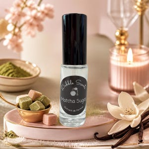 May include: A clear glass perfume roller bottle with a black cap and a circular black label. The label features the text Dabble Scents, Matcha Sugar, and Extrait de Parfum, along with a small graphic of a vintage perfume atomizer bottle.