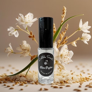 May include: A clear glass perfume bottle with a black cap, labeled "Dabble Scents Rice Papier Extrait de Parfum." The bottle is surrounded by white flowers, rice grains, and wheat stalks. The background is a soft, neutral color.