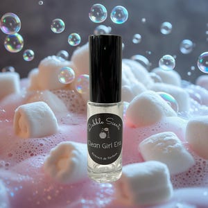 May include: A clear glass perfume bottle with a black cap, labeled "Clean Girl Era" by Dabble Scents. The bottle is surrounded by pink foam, white marshmallows, and floating bubbles. The background is a gradient of pink and gray.