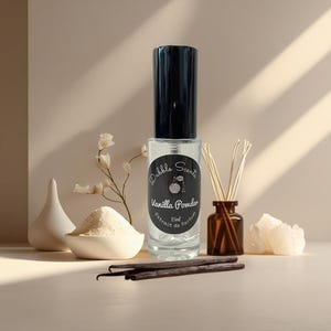 May include: A clear glass bottle of "Vanilla Powder" perfume with a black cap. The label reads "Dabble Scents". Vanilla beans, a small bowl of powder, and a diffuser are also present.