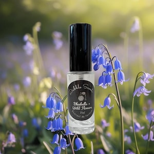 May include: A clear glass perfume bottle with a black cap, labeled "Bluebells + Wild Flowers." The bottle is surrounded by bluebell flowers, with a blurred background of more blue and purple flowers. The bottle contains 15ml of perfume.