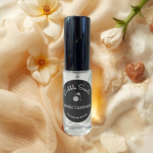 May include: A clear glass perfume bottle with a black cap, labeled "Dabble Scents Vanilla Cashmere Extrait de Parfum." The bottle is surrounded by cream-colored fabric, white flowers, and a heart-shaped decoration.