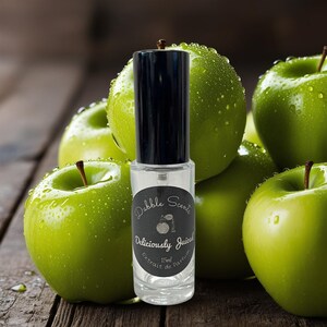May include: A clear perfume bottle with a black cap, labeled "Dabble Scents Deliciously Juiced Extrait de Parfum 15ml," is centered in front of several green apples. The apples have water droplets on them, and the background is a wooden surface.