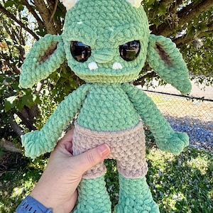 May include: A handmade crochet plush toy featuring a light green creature with large black glossy eyes, two white horns, and pointed ears. The creature has a textured yarn surface and is wearing light brown crocheted shorts with visible stitches.