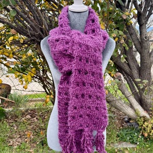 May include: A vibrant purple, hand-knitted scarf with a textured, open weave design. The scarf features a soft, fuzzy texture and decorative fringe at the ends. The scarf is displayed on a mannequin, showcasing its length and drape.