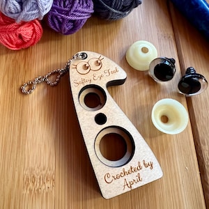 May include: A wooden safety eye tool with a chain, three different sized holes, and the text "Safety Eye Tool" and "Crocheted by April". The tool is on a wooden surface with several balls of yarn and safety eyes.