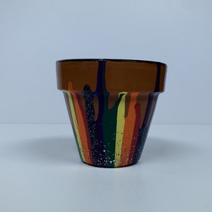 May include: A terracotta flower pot with a colorful drip paint design. The pot is painted with streaks of orange, yellow, green, blue, and purple paint. The pot has a speckled finish.