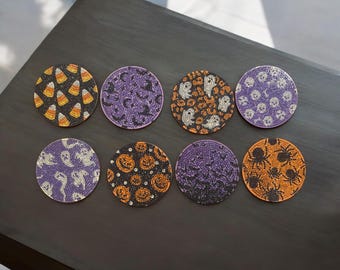 Op de afbeelding: Set van acht ronde onderzetters met Halloween-thema. Elke onderzetter heeft een ander ontwerp, waaronder snoepmaïs, vleermuizen, geesten, pompoenen, spinnen en schedels. De onderzetters zijn voornamelijk zwart, paars en oranje.