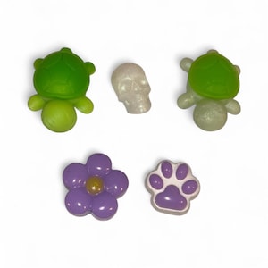 May include: A collection of decorative resin embellishments in various shapes. Includes two green turtles, a white skull, a purple flower with a yellow center, and a purple paw print.