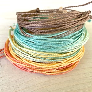 May include: A stack of colourful bracelets made from thin, braided cords. The bracelets are in shades of peach, yellow, light blue, and brown.