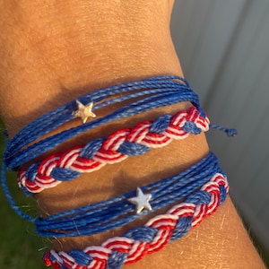 Puede incluir: Dos pulseras de amistad trenzadas en rojo, blanco y azul con abalorios de estrella plateados. Las pulseras se llevan en la muñeca.