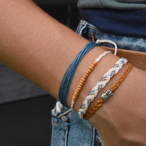 Könnte beinhalten: Ein Set aus vier Armbändern am Handgelenk einer Person. Die Armbänder sind aus blauem, weißem und braunem Kordel gefertigt. Ein Armband hat einen kleinen Ananas-Anhänger.