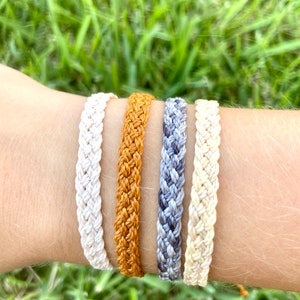 May include: Four braided friendship bracelets in white, orange, grey, and white. The bracelets are stacked on a person's wrist.