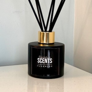 May include: Black reed diffuser with black reeds and a gold collar. The diffuser has a label that reads "Signature SCENTS HANDMADE". The product is on a white surface.