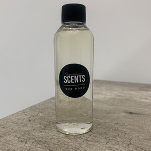 May include: A clear glass bottle with a black cap, containing a pale liquid. The bottle features a black and white label reading "Signature SCENTS OUD WOOD". The bottle rests on a grey surface.