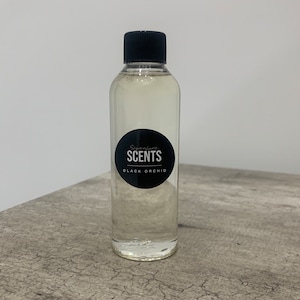 May include: A transparent glass bottle with a black lid, containing a pale liquid. A black circular label displays "Signature SCENTS BLACK ORCHID". The bottle sits on a grey, textured surface.