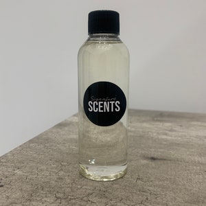 May include: A clear liquid in a transparent bottle with a black cap. The bottle has a black circular label with the words "Signature SCENTS" in white. The bottle is on a grey, textured surface.