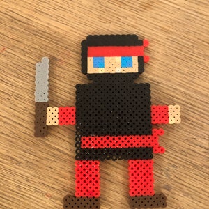 May include: A black and red pixel art figure of a ninja holding a silver knife. The ninja has a red headband and blue eyes.