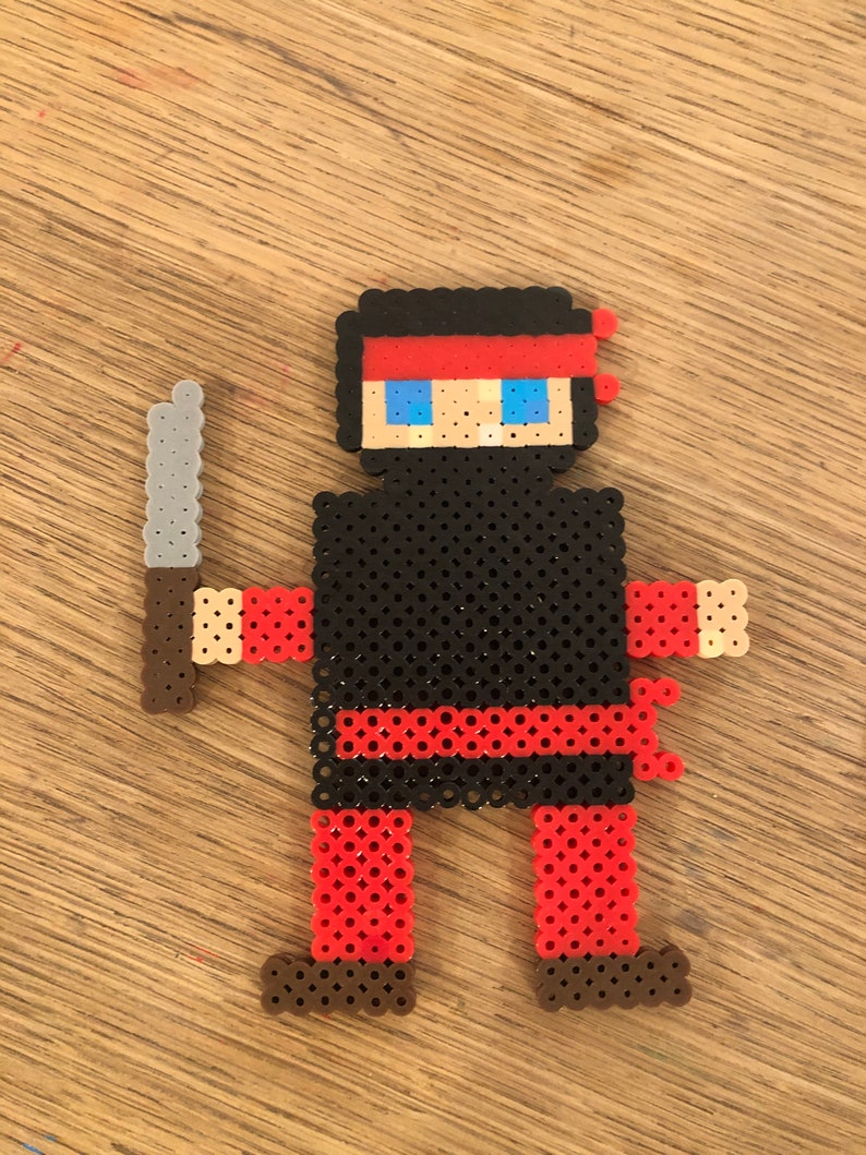 May include: A black and red pixel art figure of a ninja holding a silver knife. The ninja has a red headband and blue eyes.